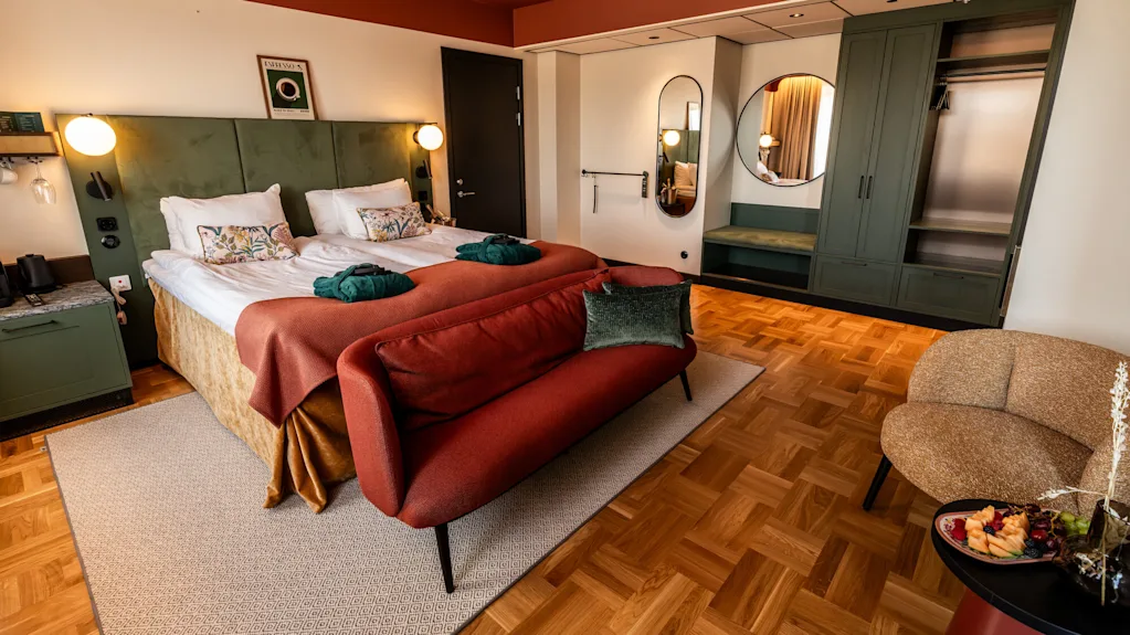 Twin beds with plush pillows and robes sit beside a green headboard. A red couch faces them, with wooden flooring and minimalist decor creating a cozy atmosphere.