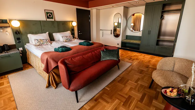 To senger med myke puter og badekåper står ved siden av et grønt sengegavl. En rød sofa vender mot dem, med tregulv og minimalistisk dekor som skaper en koselig atmosfære.