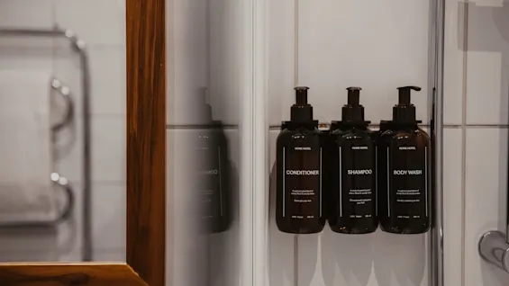 Tre mørke flasker mærket Conditioner, Shampoo og Body Wash monteret på en flisebelagt væg på et badeværelse, ved siden af et træspejl.