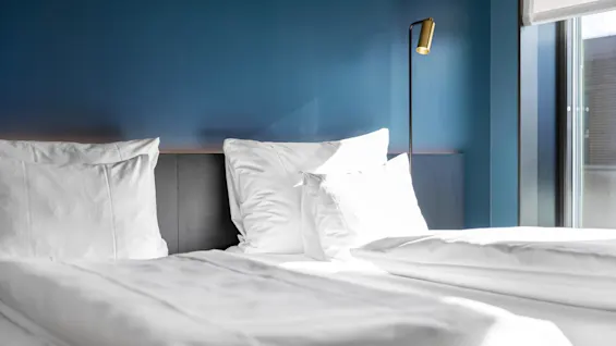 Et komfortabelt hotellrom med en seng med hvitt sengetøy, en blå aksentvegg og en moderne lampe. Naturlig lys fyller rommet.