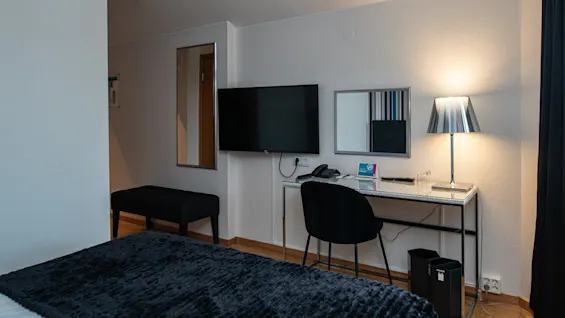 Et ryddeligt hotelværelse har et skrivebord med stol, en tændt lampe og et vægmonteret TV. En seng med mørkt sengetæppe er i forgrunden.