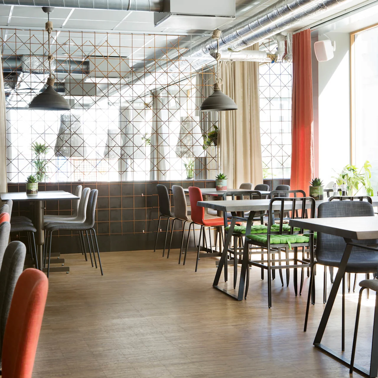 Restaurangbord och stolar står tomma, belysta av naturligt solljus genom stora fönster. Rummet är dekorerat med krukväxter, industriella lampor och jordtonade gardiner.