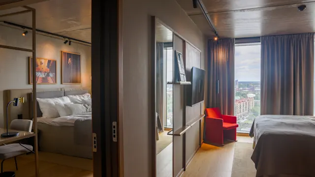 Et moderne hotelværelse med en seng mod et stort vindue, der tilbyder byudsigt. En rød stol står tæt på vinduet. Ved siden er et andet værelse med en ekstra seng.