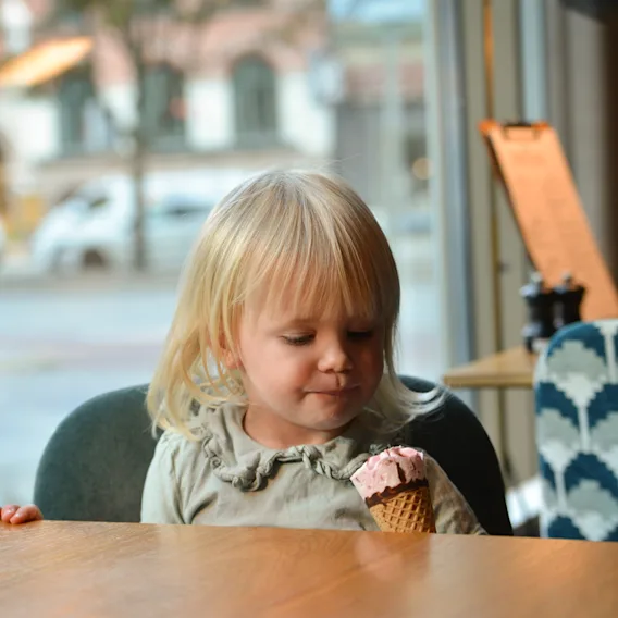 Child holding an ice cream cone, sitting at a wooden table in a cafe, with a blurred street view visible through the window.
