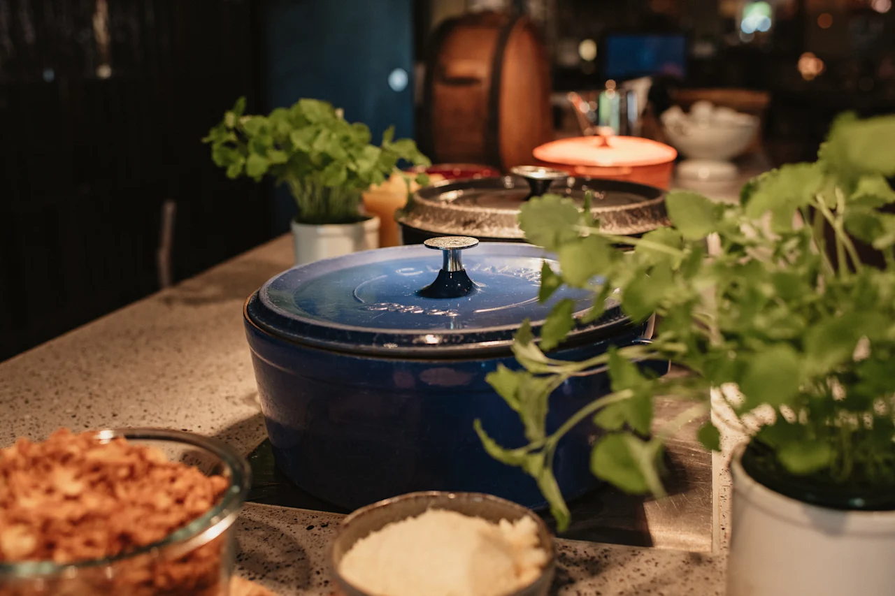 En blå kokegryte på en benk, omgitt av andre gryter, friske urter og skåler med ingredienser, klar for et måltid.