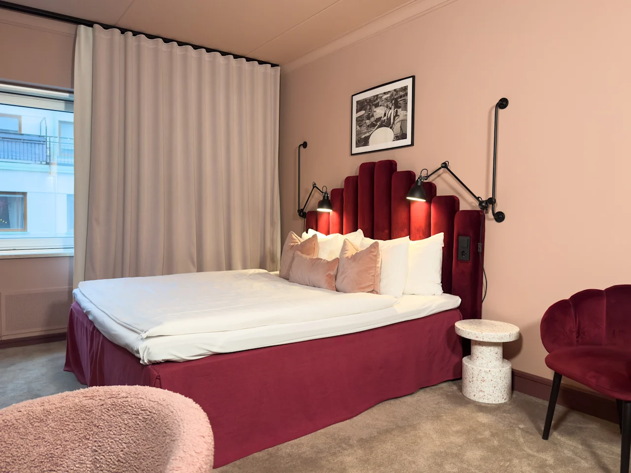 Viihtyisä hotellihuone, jossa on suuri sänky, muhkea sängynpääty ja pehmeä valaistus, kutsuen rentoutumaan ja viihtymään.