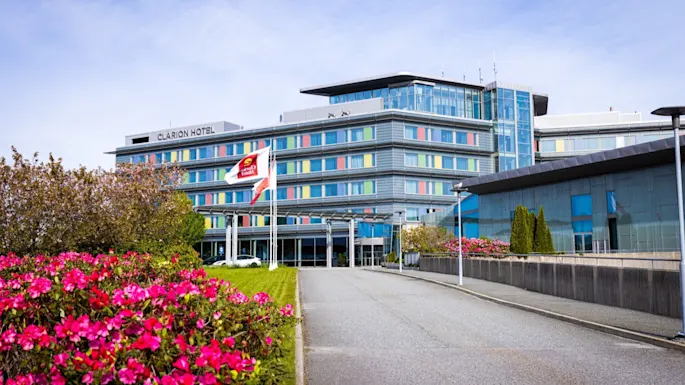 A modern, multi-story hotel labeled "CLARION HOTEL" stands amidst landscaped gardens with vibrant pink flowers. The scene is bright, under a clear blue sky. Nearby, a flag displays a hotel logo.