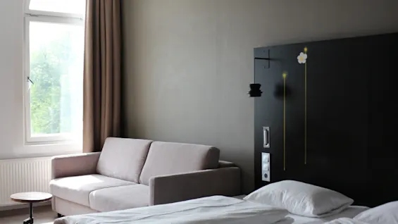 A neatly made twin bed adjacent to a gray sofa sits in a hotel room with a dark wood accent wall and an arched window revealing greenery outside.