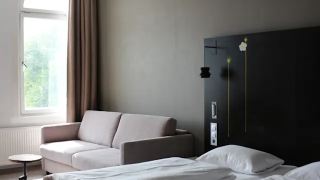 En välbäddad säng intill en grå soffa står i ett hotellrum med mörkt trävägg och ett bågformat fönster som avslöjar grönska utanför.