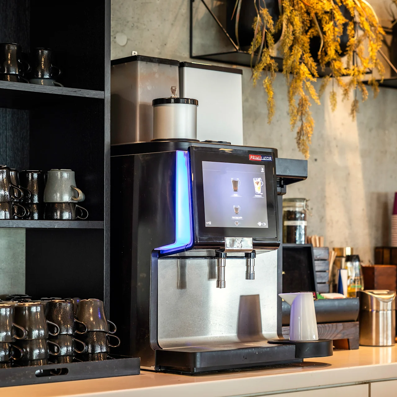 En kaffemaskin med digitala kontroller står på en disk bredvid en hylla med staplade muggar i ett café, omgivet av dekorativa växter och take away-koppar. Text: "FRANKE" på maskinen.
