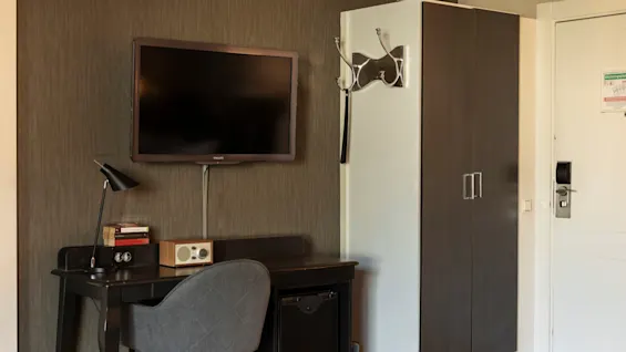 Ett modernt hotellrum med ett skrivbord med lampa och böcker, en väggmonterad TV och en garderob bredvid entrén.