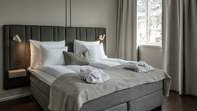 A neatly made bed with white pillows and robes sits against a gray paneled wall. Soft lighting and curtains frame a window with a view of houses.