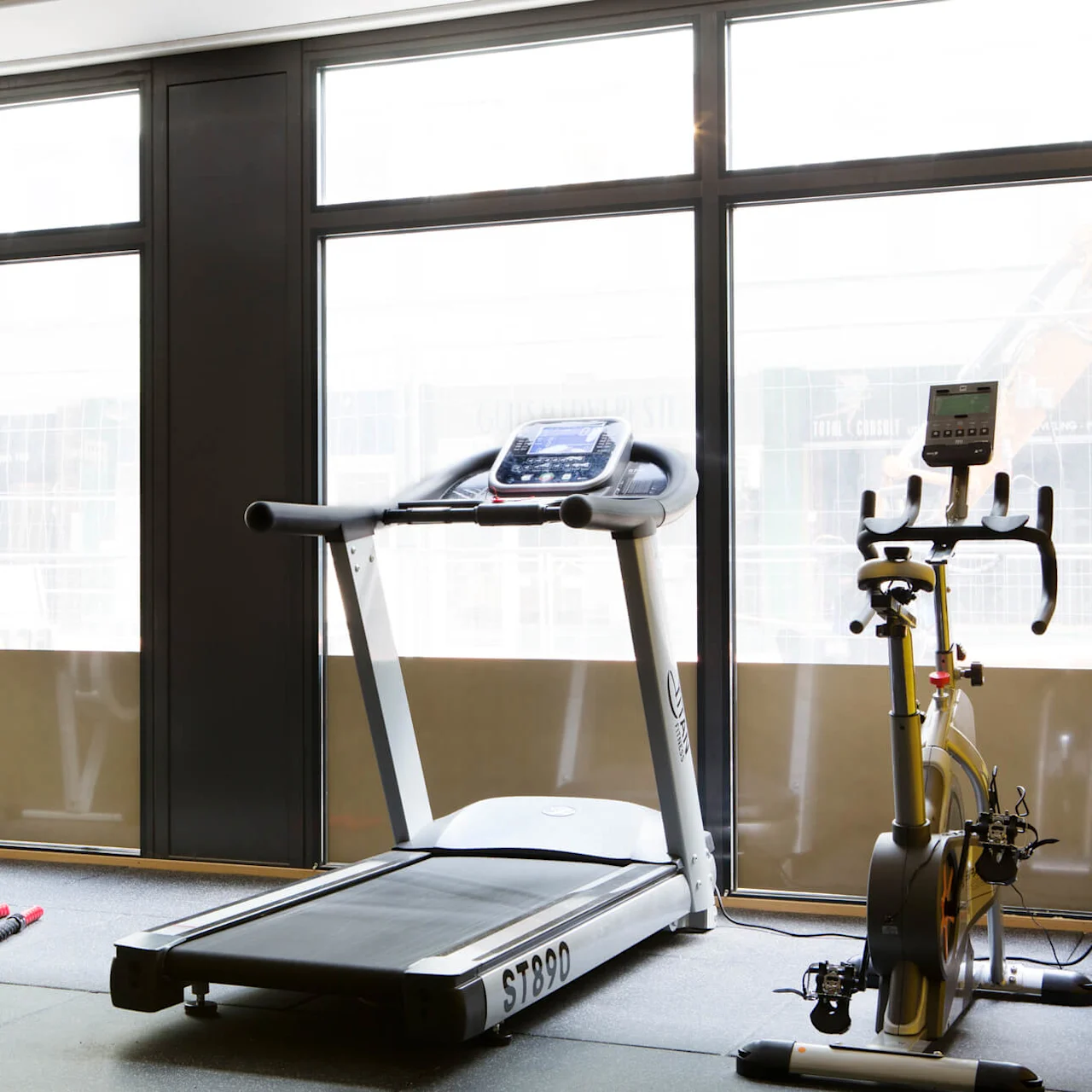 Løbebånd står stille ved siden af en stationær cykel og romaskine i et solbeskinnet fitnesscenter med store vinduer. Modelnummer på løbebåndet: ST890.