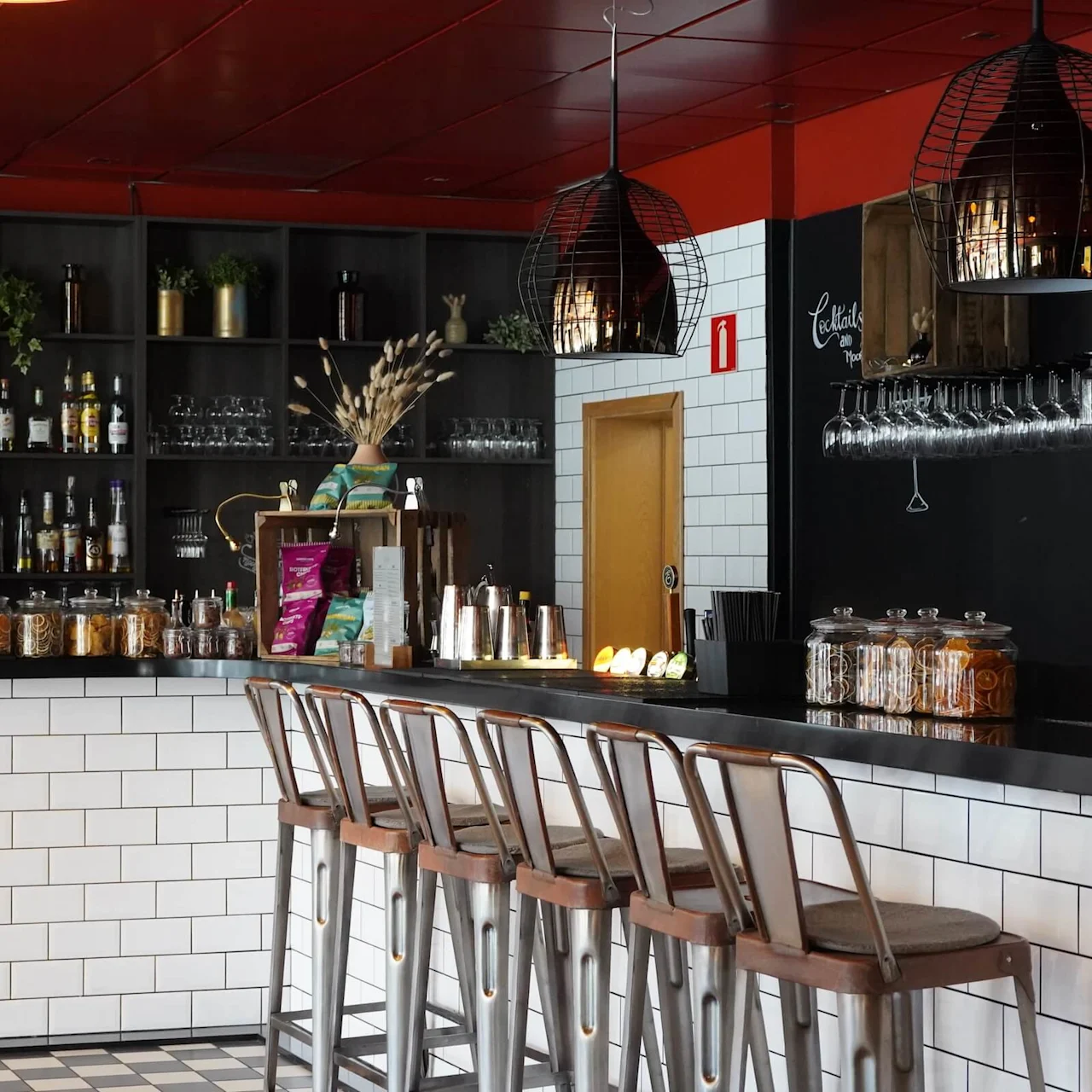 A bar with white-tiled counters displays liquor bottles on shelves, metal stools, hanging lights, and glasses. Snack jars, fruit slices, and plants add detail. Text: "Cocktails" on blackboard.