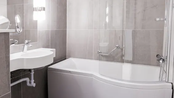 Modern badkar med en glasskärm bredvid en elegant vit vask. Ljusstark belysning och grå kaklade väggar skapar en ren, modern badrumsmiljö.