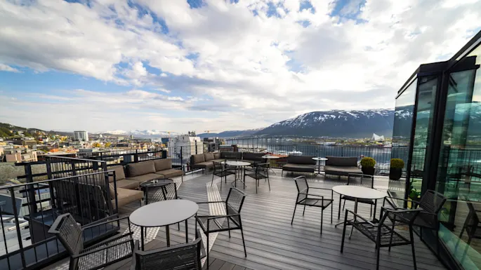Outdoor terrace with empty tables and chairs, framed by glass panels, overlooks a cityscape with colorful buildings, a body of water, and snowcapped mountains under a partly cloudy sky.