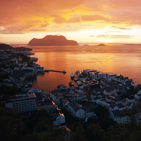 Naturskjønn utsikt over Ålesund by og havn i solnedgang, med gyllenoransje himmel og sjø.