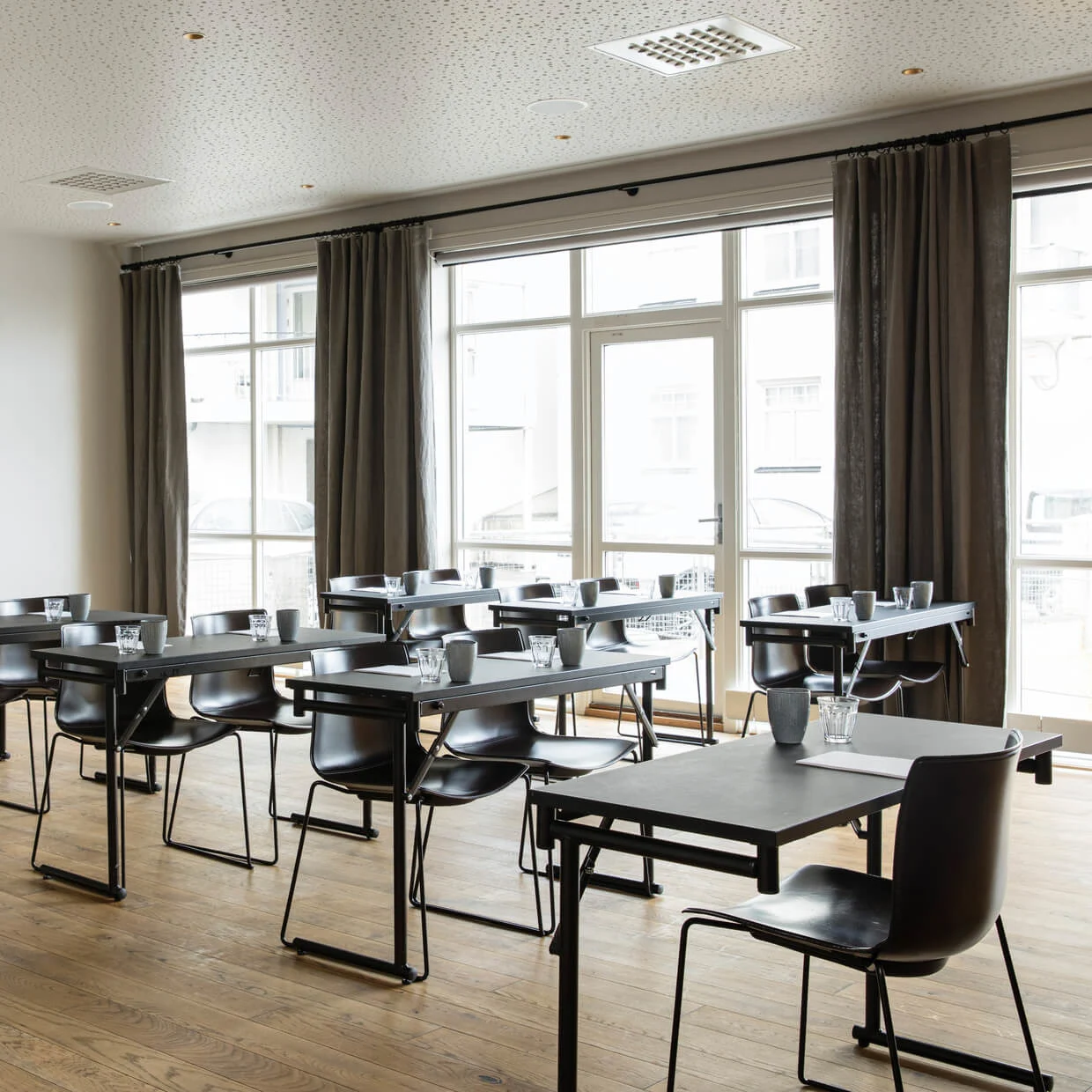 Tables are arranged in rows with chairs, glasses, cups, and papers on them. Large windows with curtains provide light. A colorful painting decorates the wall.