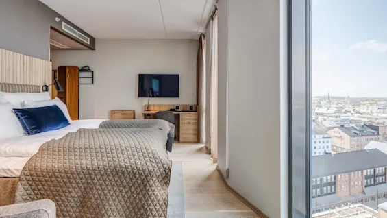 A bed with a textured blanket and blue cushion stands in a modern hotel room. A flat-screen TV is mounted on the wall opposite, next to a large window with a city view.