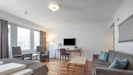 En stue har grå sofaer og lænestole, et sofabord i træ og et vægmonteret fjernsyn. Store vinduer giver naturligt lys, sammen med gardiner og en standerlampe.
