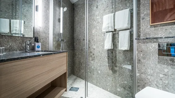 A modern bathroom features a glass-enclosed shower with two white towels hanging. A wooden vanity with a sink and large mirror is positioned nearby, on a stone-tiled wall.