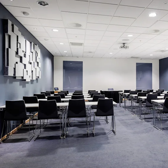 Rader av svarte stoler og hvite bord er arrangert klasseromstil i et lyst konferanserom med geometriske akustiske paneler på marineblå vegger.