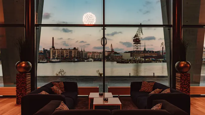 En moderne lounge med mørke sofaer og mønstrede puter vender mot et stort vindu, som viser en havneutsikt med bygninger, kraner og en solnedgangshimmel, mens fyrverkeri lyser opp horisonten.