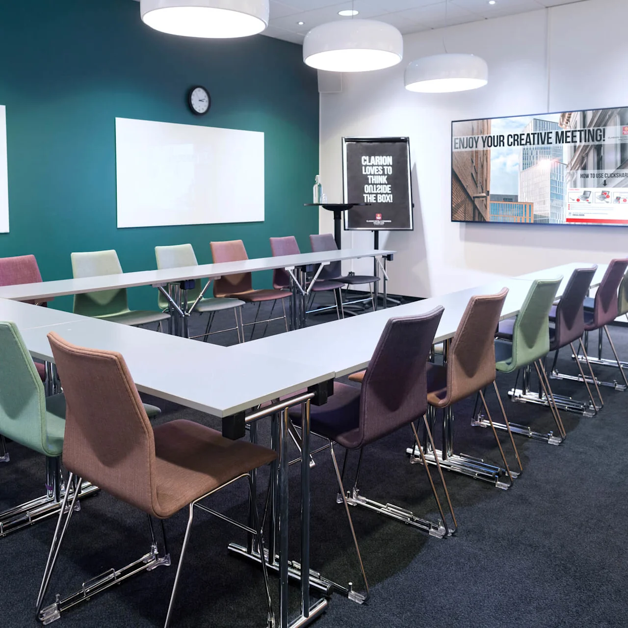Bord danner en U-formet arrangering omringet av fargerike stoler i et konferanserom med grønne vegger. "CLARION LOVES TO THINK OUTSIDE THE BOX" og "ENJOY YOUR CREATIVE MEETING!" på tavlen.
