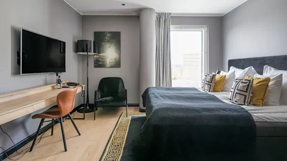 A modern bedroom features a double bed with colorful cushions, a mounted TV, a desk with a chair underneath, artwork on the wall, and a window with light curtains.