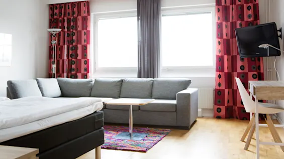 A gray sofa sits beside a wall of windows, under red patterned curtains. A bed and a small wooden table are nearby, in a brightly lit room with hardwood floors.