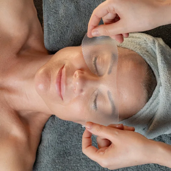 A person receiving a facial treatment, with a mesh mask being applied over their eyes. They are wrapped in a towel in a spa setting.