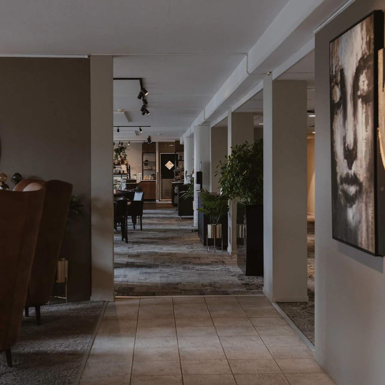 A hallway displays modern art on the wall, featuring abstract faces, leading to a dining area. Soft lighting and plants create a cozy atmosphere.