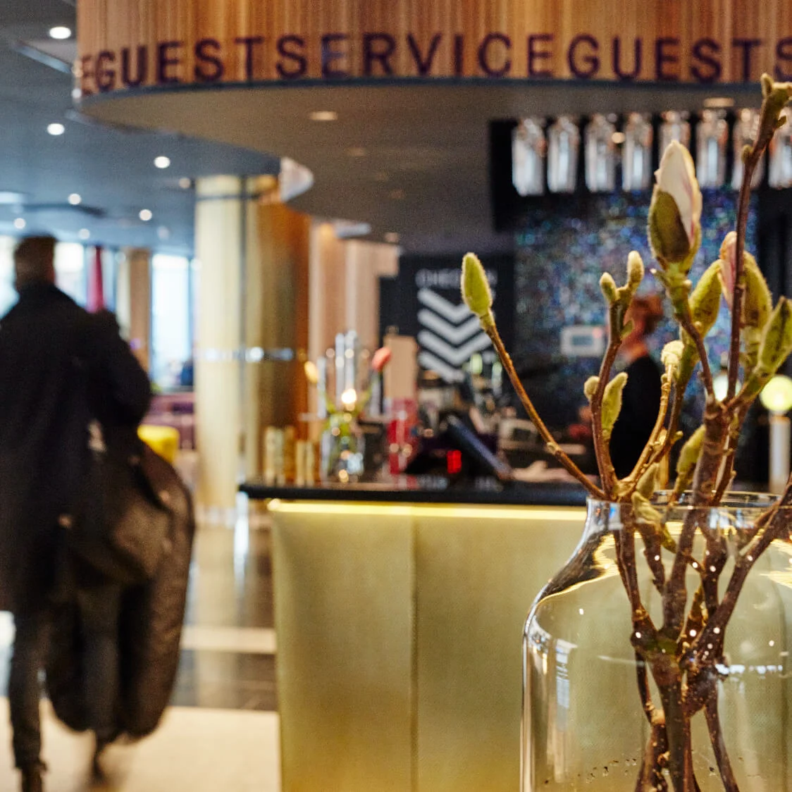 En person går gennem en stilfuld hotellobby og trækker en kuffert. Det moderne rum har gyldne søjler, lilla siddepladser og en reception med blomsterarrangement. Tekst: "GUEST SERVICE".