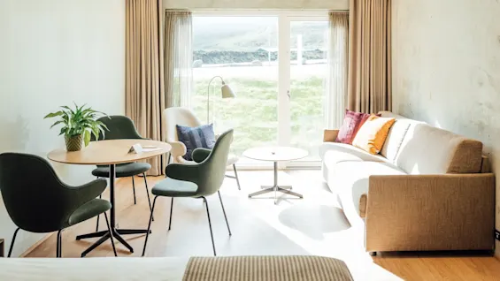 En beige sofa med farverige puder står i et lyst rum overfor et stort vindue. I nærheden er et lille bord og stole med en plante, der tilføjer grønt.
