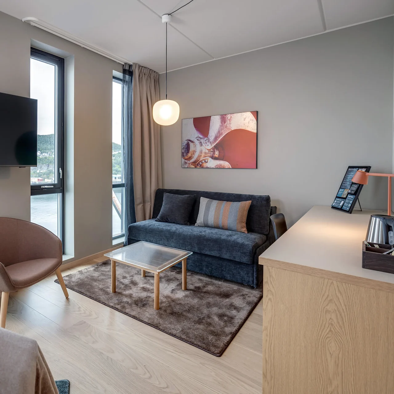 Et hyggeligt hotelværelse har en sofa og lænestol ved gulv-til-loft-vinduer. Et vægmonteret TV, et bord og et skrivebord med lampe fuldender den moderne indretning.