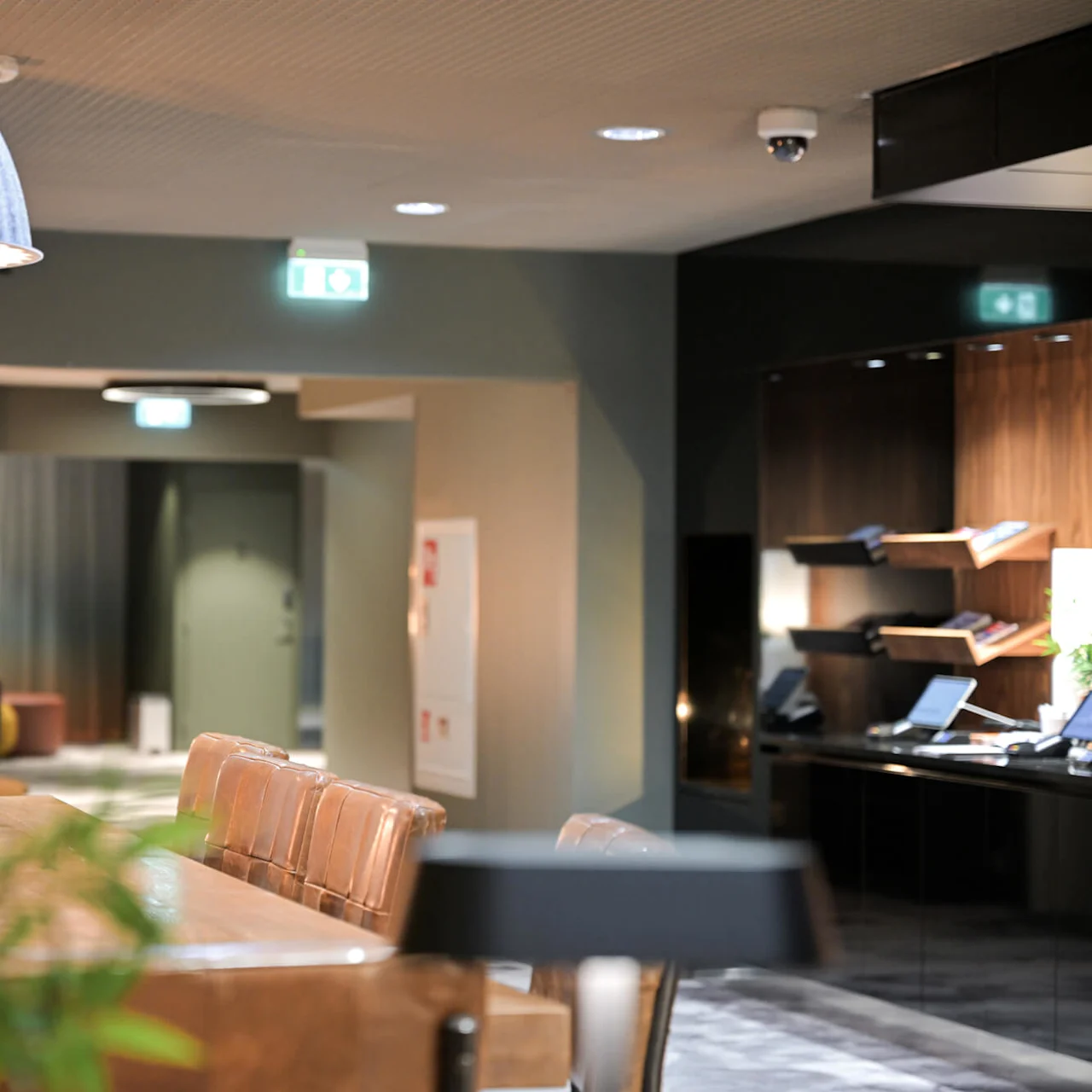 En stor, grå lampskärm lyser upp ett stilfullt rum med läderstolar och ett träbord. I bakgrunden leder en korridor till olika utgångar, markerade av lysande skyltar.