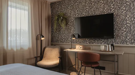 A cozy hotel room with a floral wallpaper, featuring a desk area with a TV, a comfortable chair, and a window.