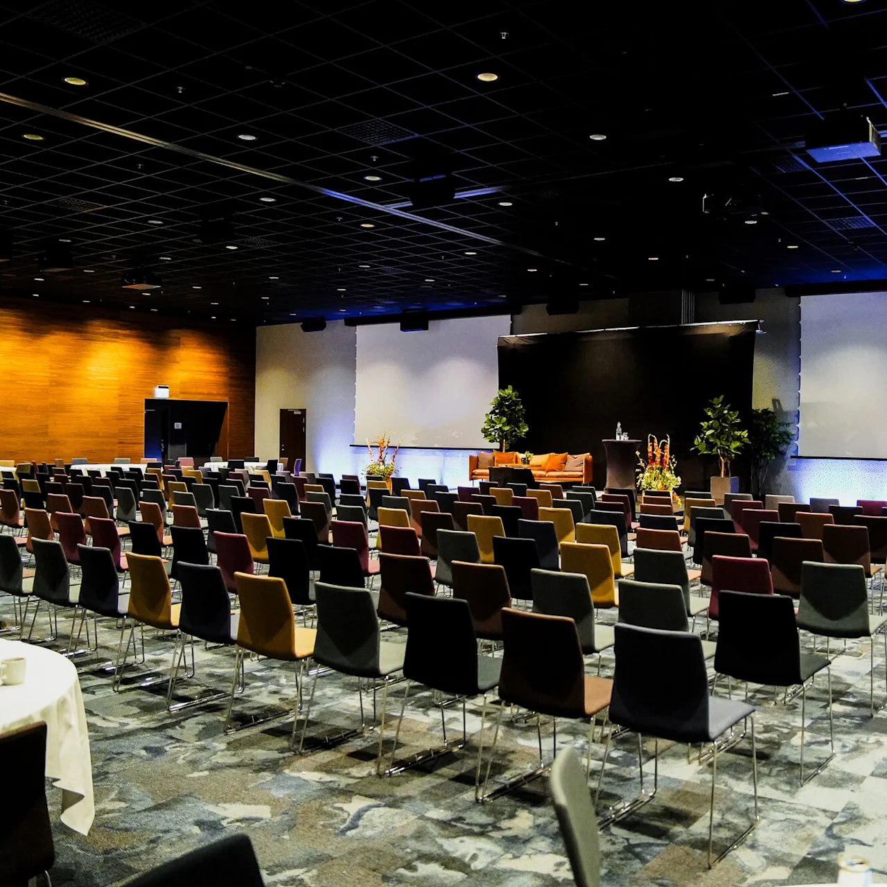 Rækker af farverige stole vender mod en stor skærm i et dæmpet oplyst konferencerum. Scenen er pyntet med planter og podie, hvilket skaber en formel præsentationsindstilling.
