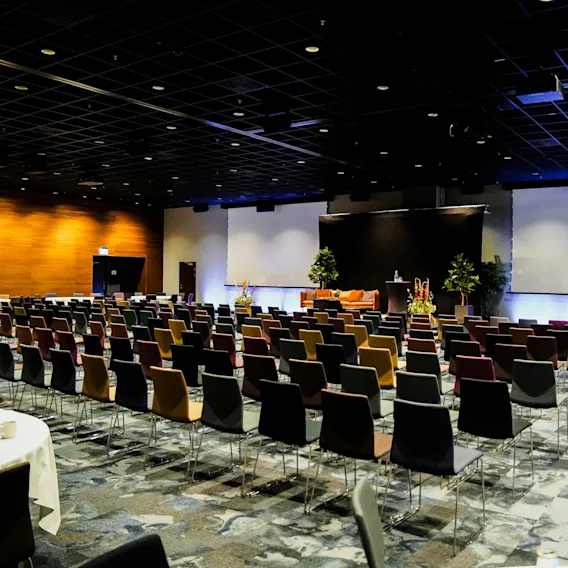 Rader av färgglada stolar är vända mot en stor skärm i ett svagt belyst konferensrum. Scenen är utsmyckad med växter och podier, vilket skapar en formell presentationsmiljö.