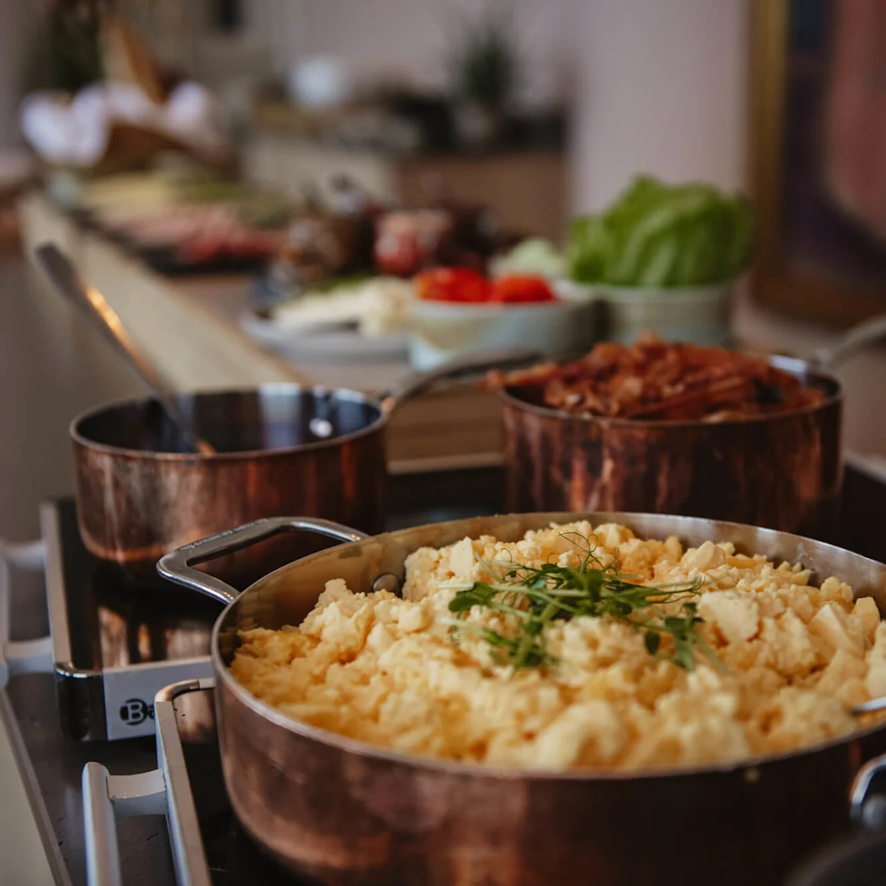Scrambled eggs in a copper pan, garnished with herbs. Nearby are pans with bacon and sausages. A buffet table is in the background with various breakfast items.