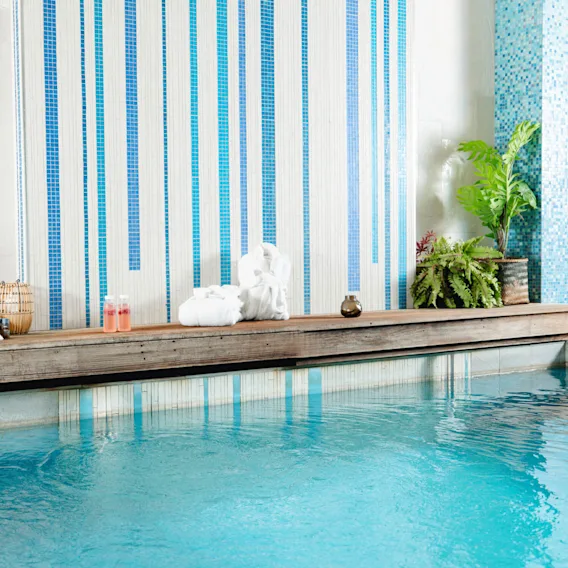 Pool omgiven av ett trädäck med handdukar, växter och två rosa drycker. Vertikala blå och vita plattor skapar en lugnande bakgrund. Spa-liknande miljö.
