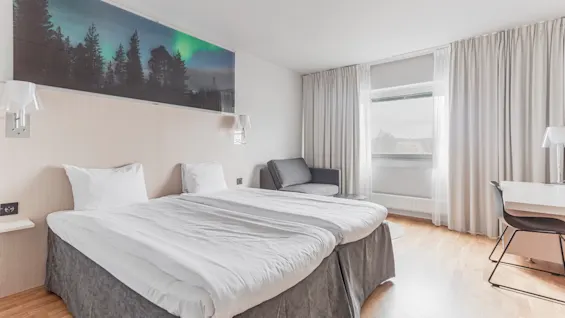 A bright hotel room with two beds, a Northern Lights art piece above the headboard, a cozy armchair, and a desk by the window.