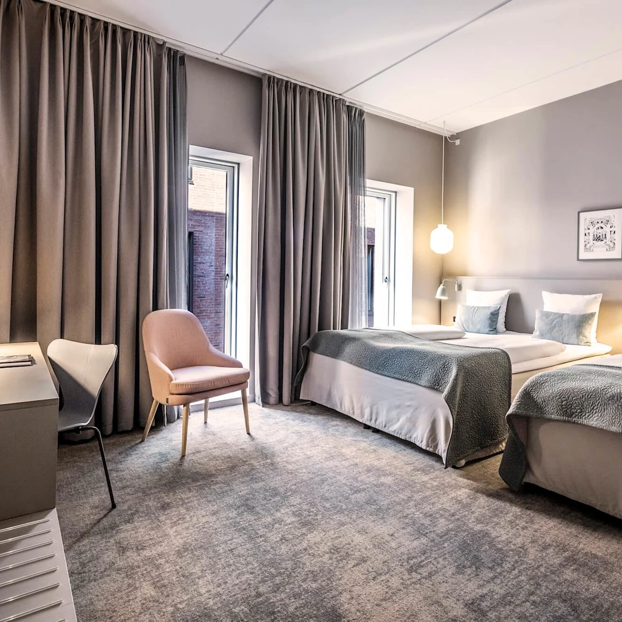 Kaksi siististi pedattua sänkyä on modernissa hotellihuoneessa, ympärillä pehmeä valaistus, pöytä, tuolit ja lattiasta kattoon ulottuvat verhot, luoden viihtyisän ja kutsuvan ilmapiirin.