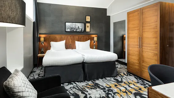 A modern hotel room with twin beds, wooden furniture, and monochromatic decor.