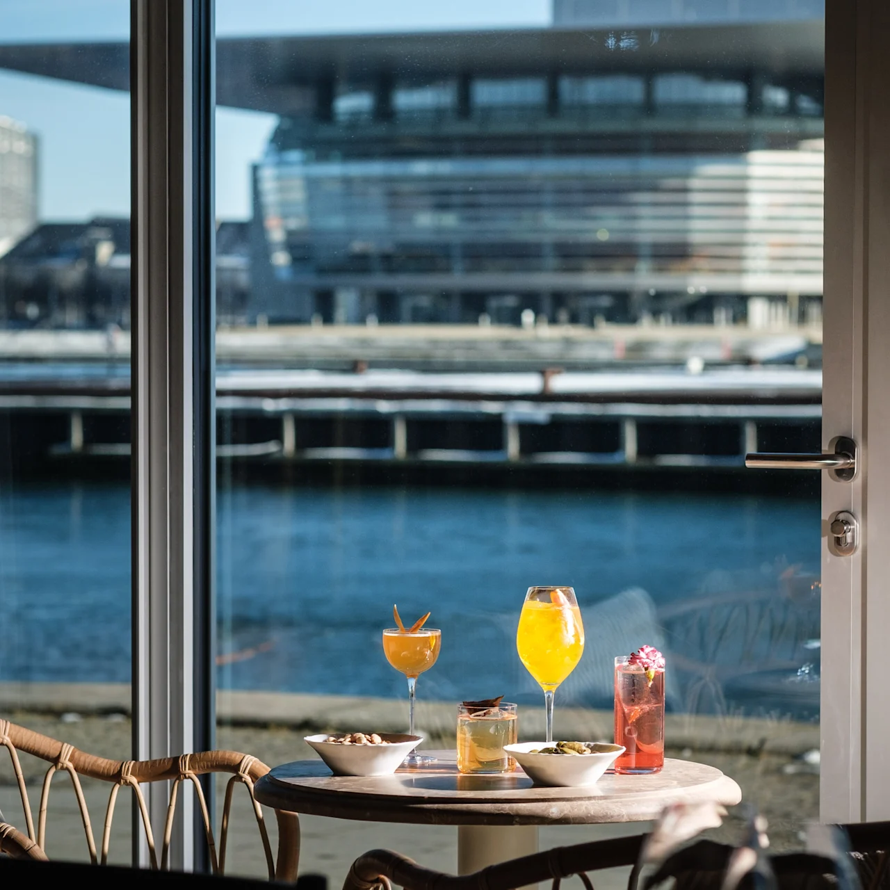 Table with three colorful drinks and snacks sits by a window, overlooking a waterfront with a modern building in the background under a clear blue sky.
