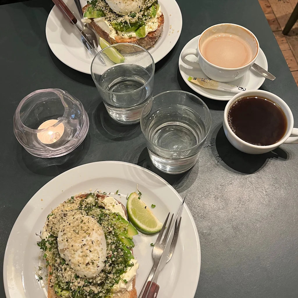 To tallerkener med avocadotoast, pocherede æg og limebåde, ledsaget af kaffe, latte og vand, dækket på et bord med et lys.