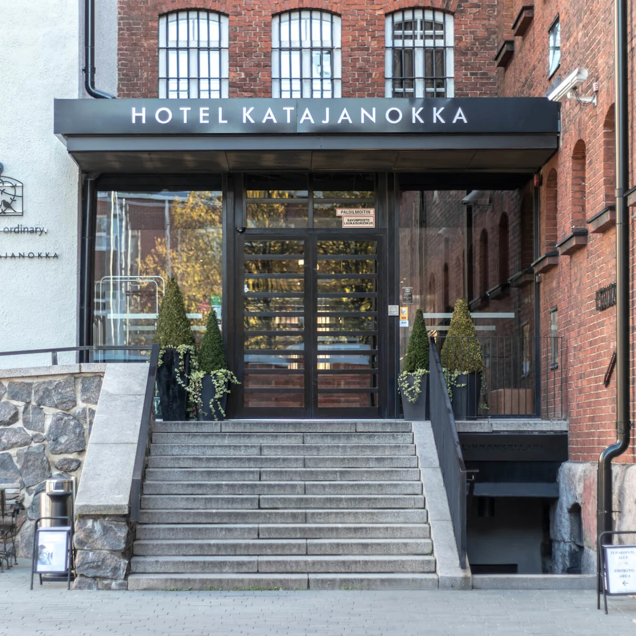 Red-brick building entrance with 'HOTEL KATAJANOKKA' sign, stone stairs leading up to glass doors surrounded by potted plants.