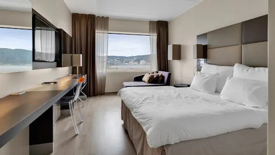 Modern hotel room features a neatly made bed with white linens beside a window overlooking a cityscape. A desk and chair are near a wall-mounted TV, surrounded by warm, neutral decor.