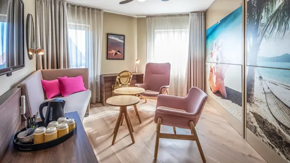 A cozy living room with a wooden table and chairs, featuring warm-toned furniture. Pink pillows adorn the sofa. Windows with beige curtains let in natural light. Large wall photo depicts a beach scene.