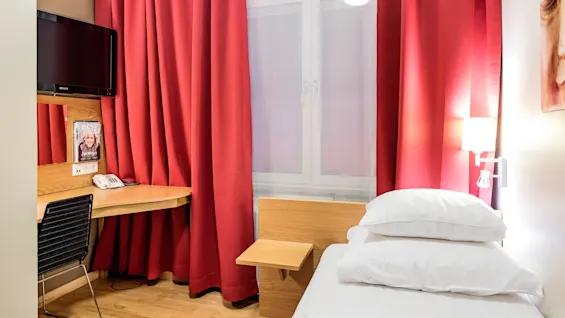 Et lite hotellrom inneholder en enkeltseng med hvitt sengetøy, en veggmontert TV over et skrivebord av tre, og røde gardiner ved vinduet. En telefon og magasinet "BEST HOLIDAY" hviler på skrivebordet.